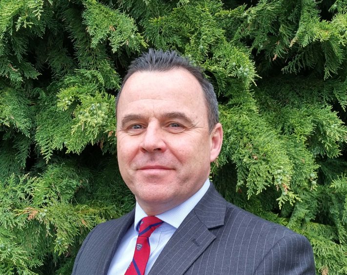 Steve Walsh, smiling at the camera. mr Walsh is in a grey pinstriped suit with a tree behind him, He is wearing a red and blue tie. 
