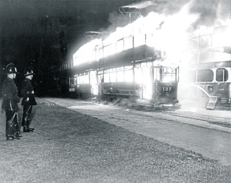Firefighters on standby stand by and watch as the intense conflagration consumes the trams.