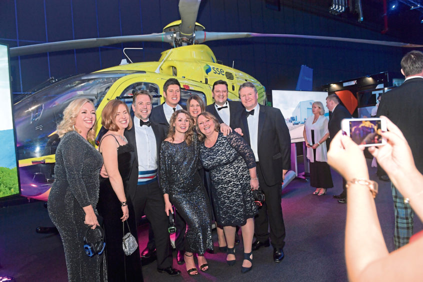 A group of people in formal evening wear posing for a photo in front of a helicopter at the event.