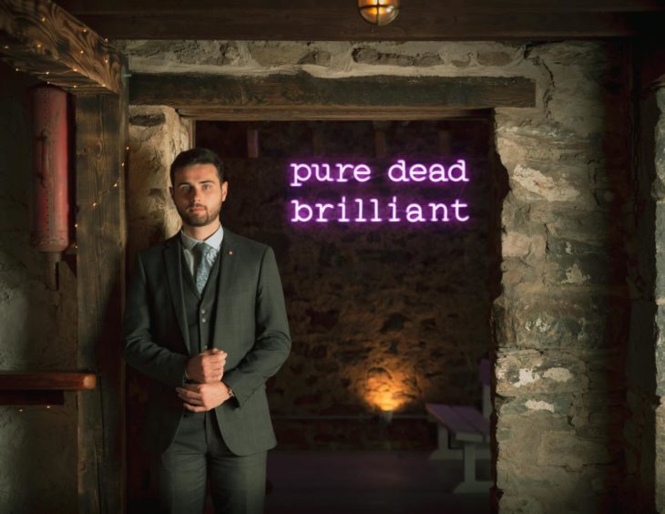 A man in a suit standing next to a neon sign that reads: 'pure dead brilliant'