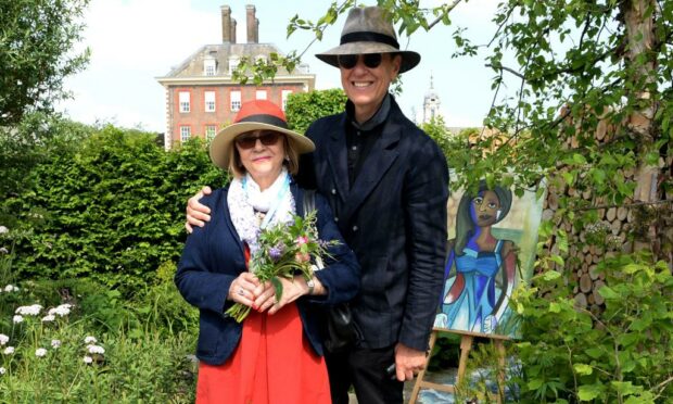 Joan Washington and Richard E Grant standing side by side wearing sun hats and sunglasses in front of greenery in London