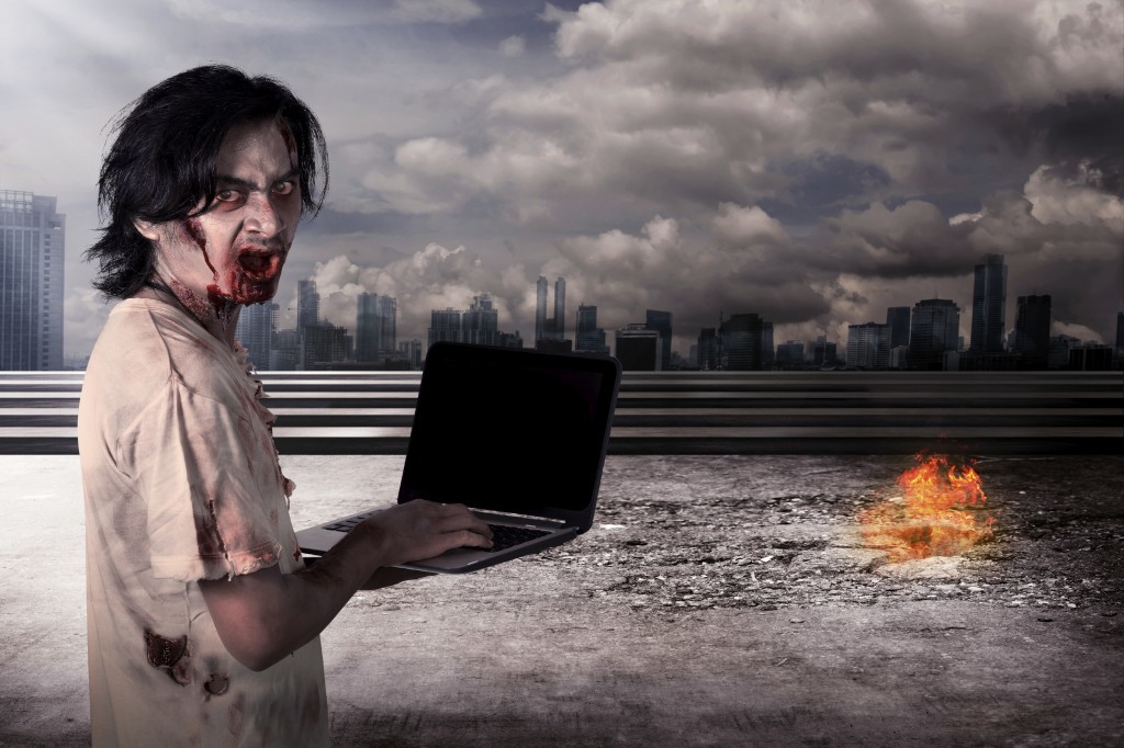 Creepy male zombie typing with laptop with city on fire background