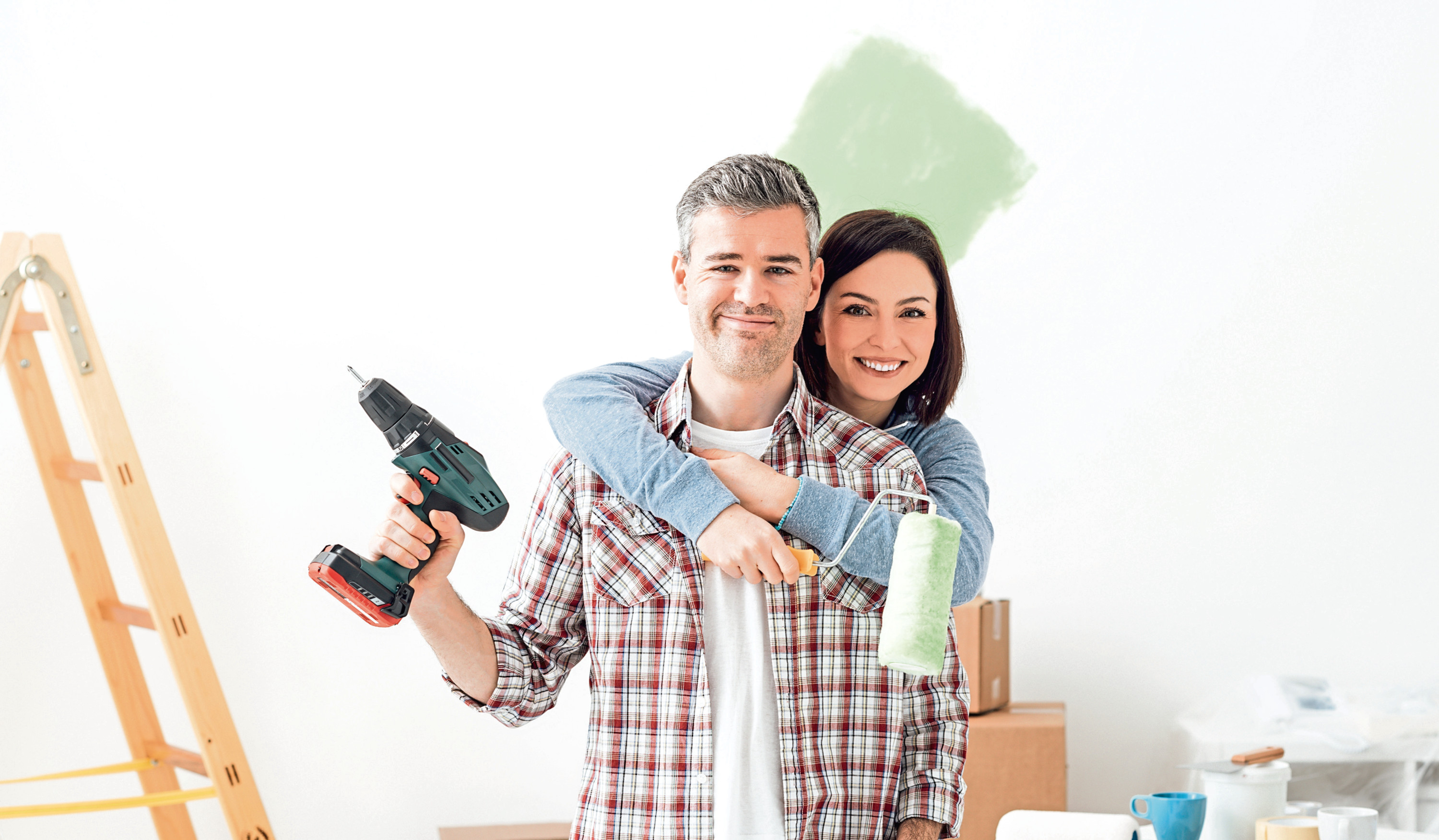 Smiling loving couple doing home renovations, the woman is holding a paint roller and the man is using a drill