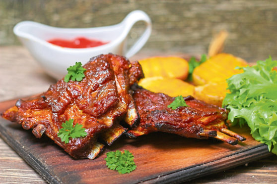 Appetizing rosy and barbecued lamb ribs seasoned with a barbecue sauce and served with fresh herbs and potatoes on an old rustic wooden chopping board