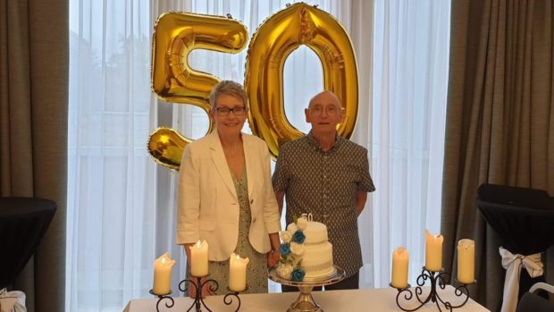 Joe and Doris Boag stand in front of two gold ballons (one 5 and one 0), behind a cake, celebrating their 50th wedding anniversary.