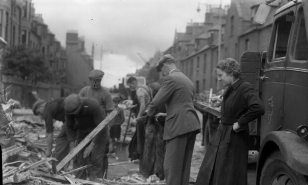 A black and white image of men clearing rubble following the Peterhead bombing. A woman looks on, hands on hips, in a dark coat as the suited men, all wearing flat caps search through piles of wood and rubble.