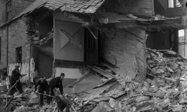 A black and white picture showing the gable end of a house completely destroyed by the Peterhead bombing. Three men in suits and hats shovel debris.