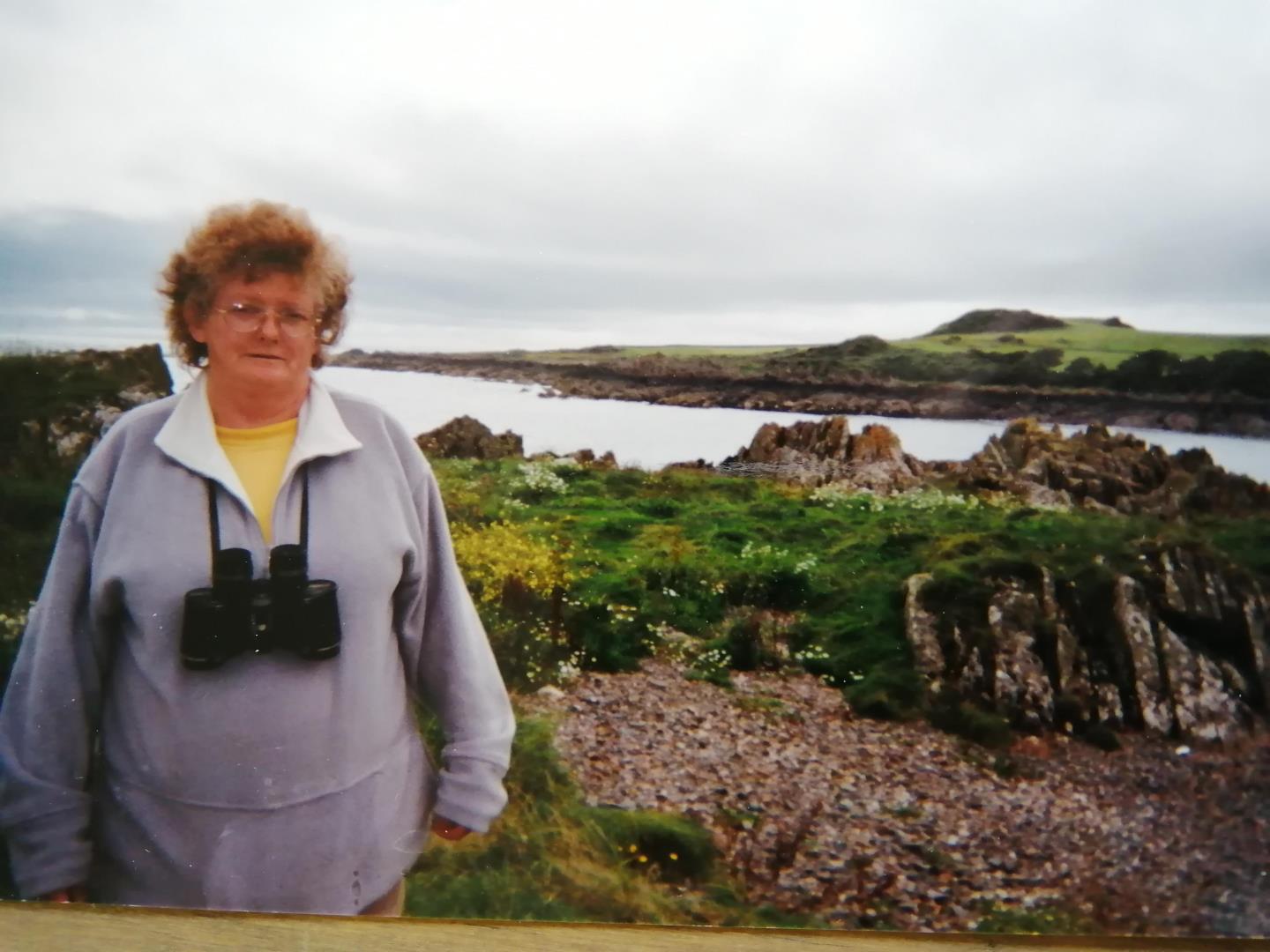 Joan Humphreys stands near the coast wearing binoculars