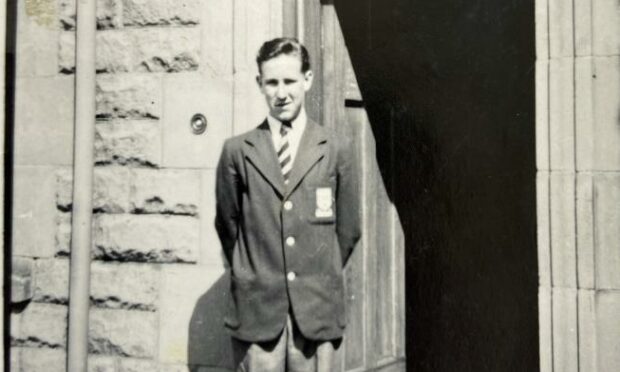 A young Ken Gordon is shown at the stone doorway of George Heriot school. He is wearing a striped school tie and badged blazer. 