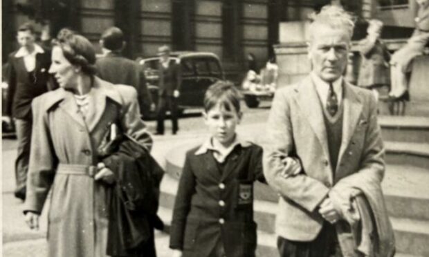 A black and white picture showing a young Ken Gordon holding the arm of his father, while his mother walks behind them on a busy Aberdeen street. Black cars and crowds are also shown.