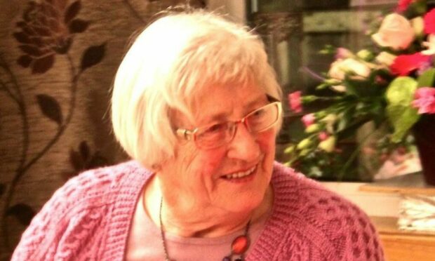 Smiling in an unposed way, Evie MacKenzie is pictured wearing a pink knitted cardigan, pink t-shirt and multi-coloured necklace. She has flowers behind her and she has glasses on.