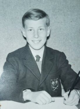 A black and white image shows a young, smiling Iain McIntosh sitting at a desk with a pencil and paper, wearing a badged school blazer.