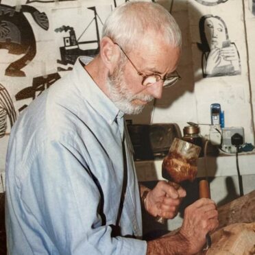 Coloured image depicting Iain McIntosh, wearing glasses with white hair, carving wood. he is pictured in front of a patterned back drop.