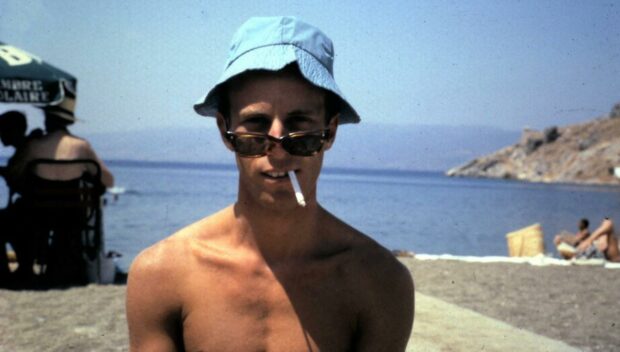 Shown during a trip to Greece, artist Iain McIntosh is pictured in his student days shirtless, wearing a blue bucket hat, sunglasses low on his nose, with a cigarette hanging from his mouth. The sea and coastline is shown behind him.