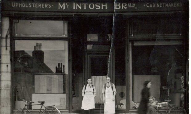 James and Bill McIntosh are pictured in a black and white shot, outside the shop they owned in Peterhead. They are in front of a high-ceilinged and large windowed shop exterior wearing white aprons over shirts and ties. 