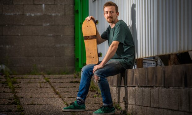 Malcolm Bradley sitting on a brick wall showing off a small skateboard made from cork and wood.