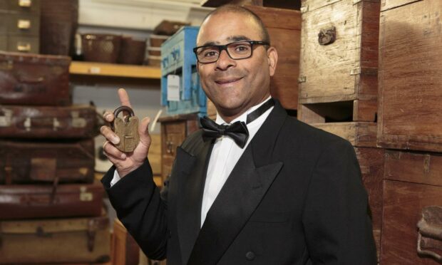 Carl Morenikeji is wearing a James Bond-like suit holding up a large vintage brass padlock.