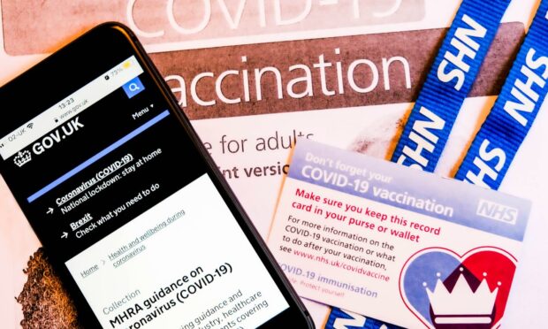 A mobile phone showing MHRA guidance, alongside an NHS lanyard and Covid vaccination card/.