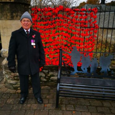 Michael Cheesewright is shown in front of a bright red poppy display wearing his medals 