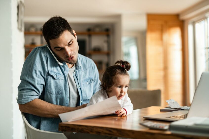 A man working from home with a phone to his ear, papers in his hand and a toddler in his lap, looking like he could use some advice for working when schools are closed