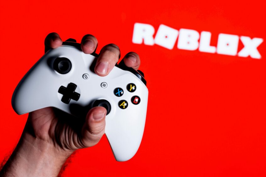 A hand holding a games console controller with the Roblox logo in the background.