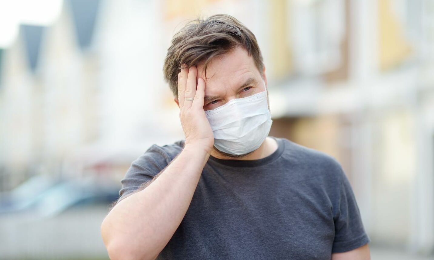 A man wearing a face mask stands in the street, holding the side of his head