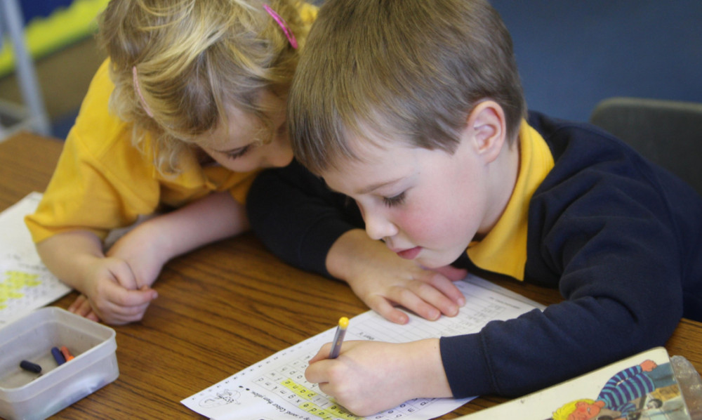 two children writing with a pencil and paper at a desk.