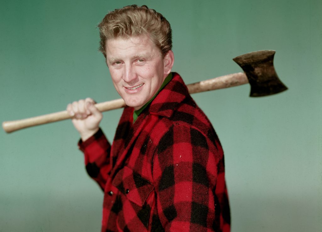 Portrait of American actor Kirk Douglas wearing a black and red checked jacket and holding an axe over his shoulder, 1950s. 