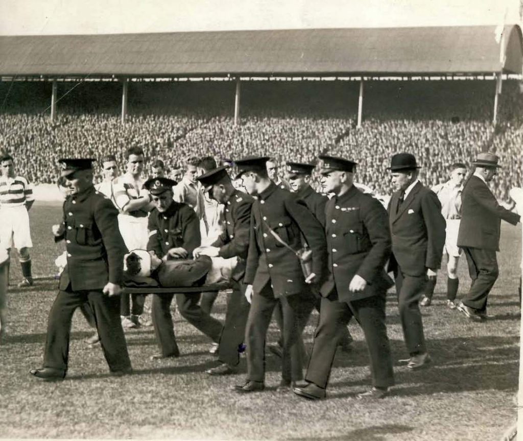 Thomson being carried off the pitch after the collision, followed by Bill Struth, Rangers manager. Willie Maley, Celtic manager is on the extreme right.