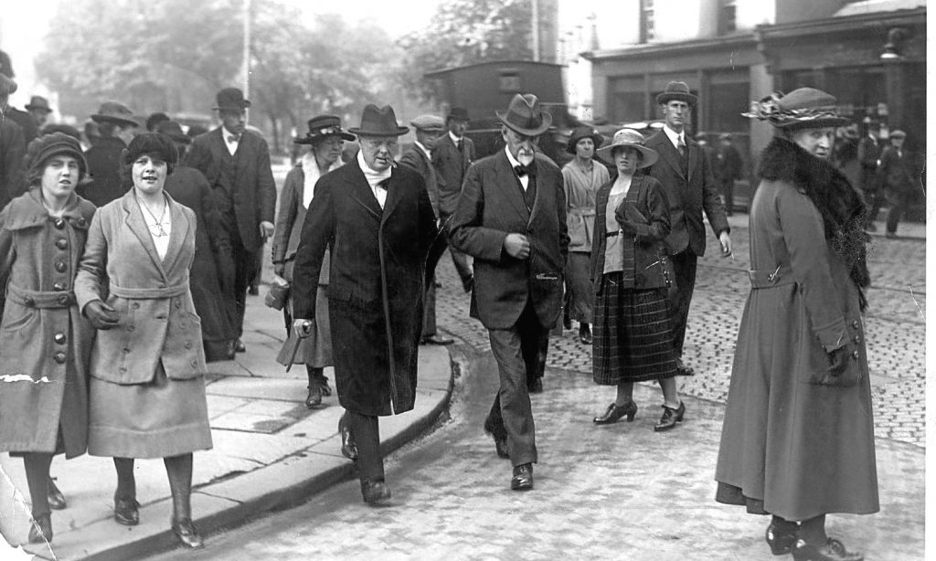 Black and white photo of Winston Churchill in long dark overcoat and hat on a busy street in Dundee in the early part of the 1900s.