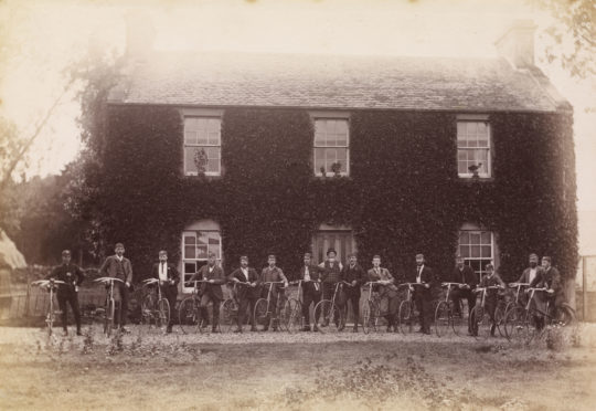 View of group of fifteen men with bicycles outside an ivy-clad house, possibly in the Stanley area.