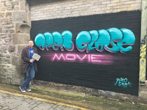 Jon Gill with a mural depicting the logo for his documentary, painted in an alleyway near the Dundee bus station.