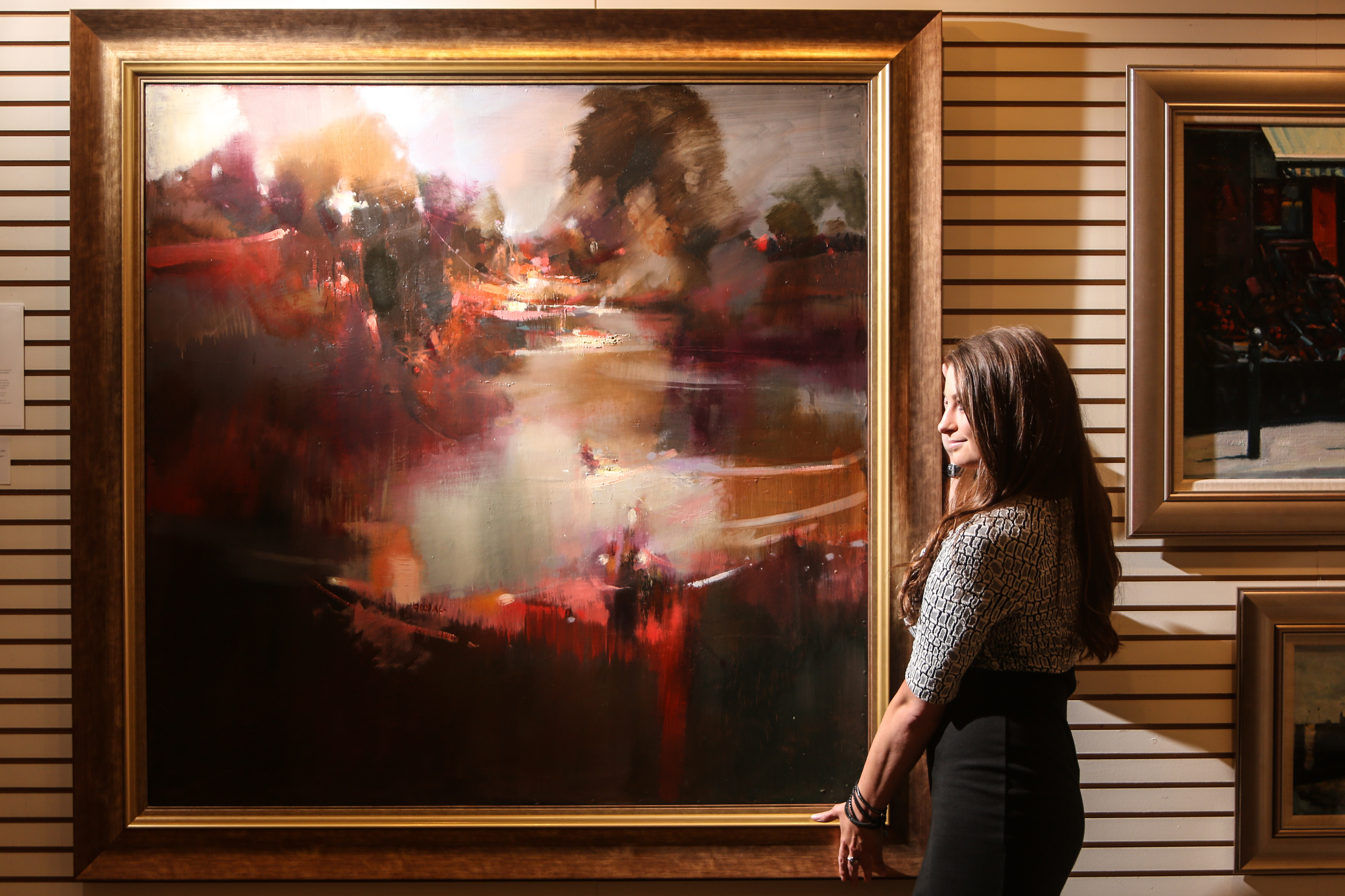 Kate Cunningham, gallery assistant and artist with a painting by Oscar Goodall RSW, entitled Weir and River.