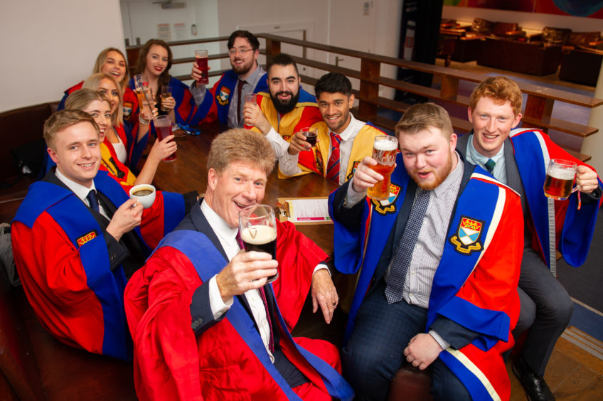 Jim Spence, in rector robes, raising a pint of Guinness at a table with Dundee University students.