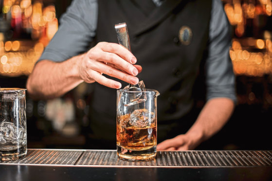 Bartender adding a big ice cube to the measuring glass cup with a brown cocktail on the bar counter on the blurred background; Shutterstock ID 1361183939; Purchase Order: -