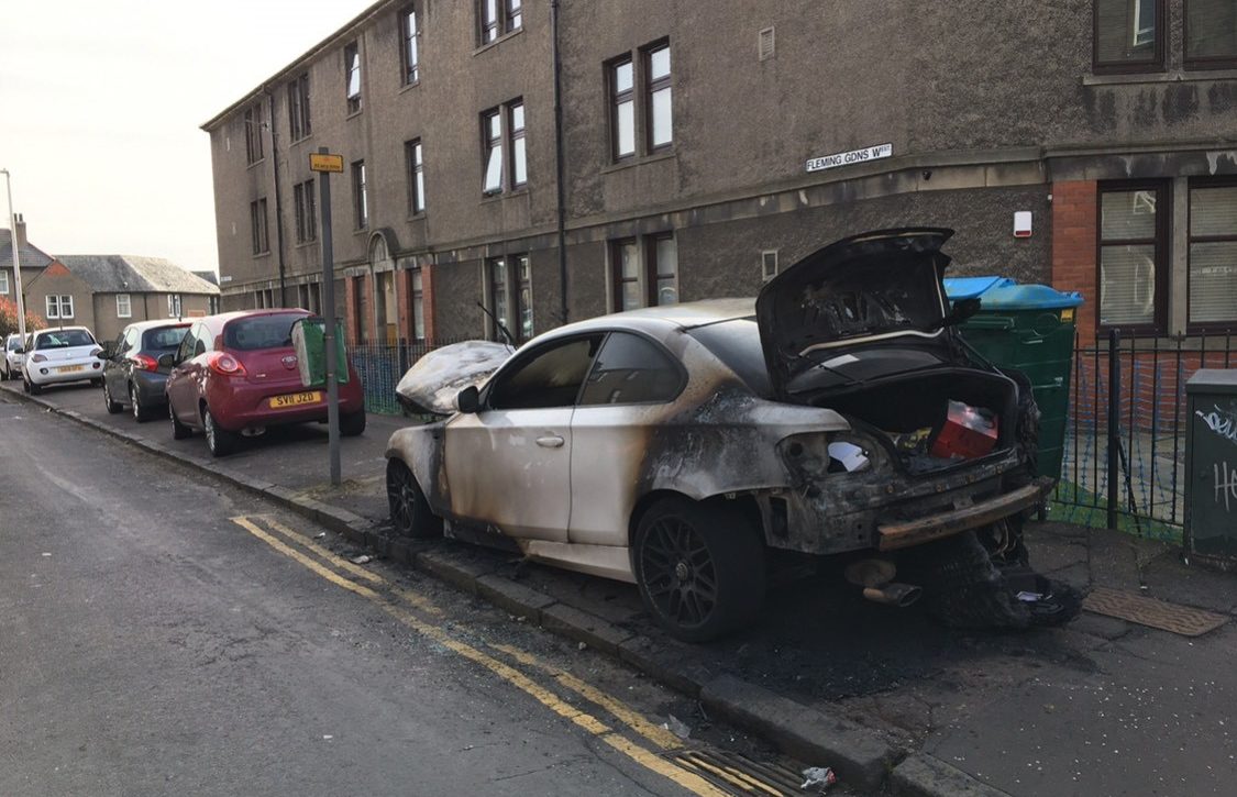 A row of cars parked on the pavement, alongside one that was burned.