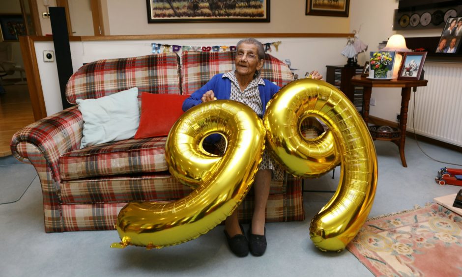 Daphne Shah seated on sofa with two large 9 balloons for her 99th birthday