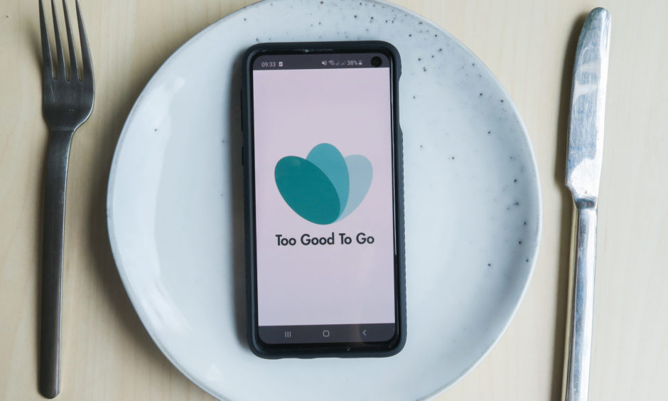 The Too Good to Go logo on a phone resting on a plate with knife and fork by the side