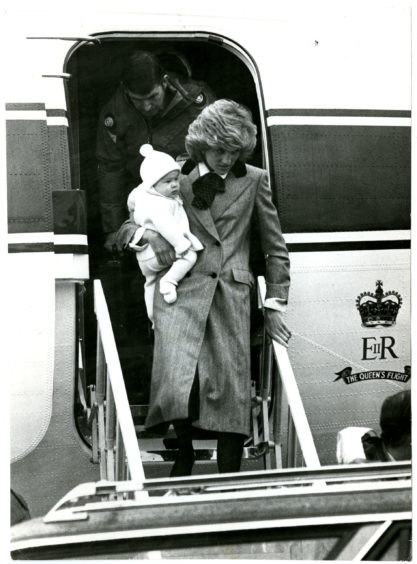 Black and white photo shows Princess Diana getting off a flight with the Queen's crest on the side, carrying the infant Prince Harry in 1985.