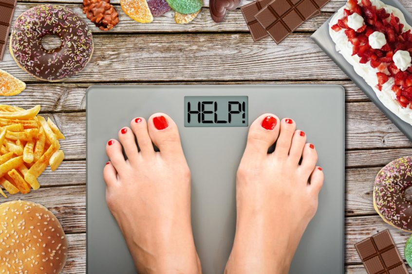 woman on scales, which spell out the word 'HELP', surrounded by junk food.