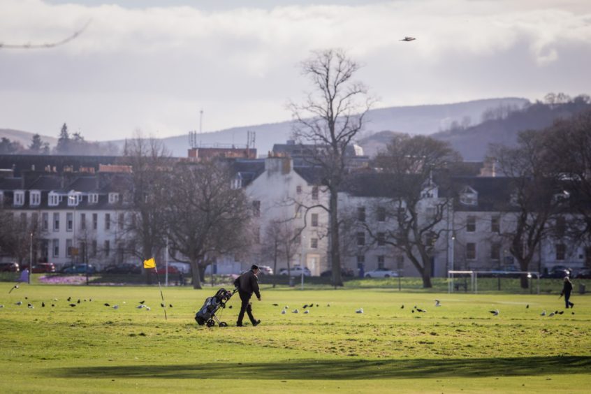 Golfer walking on North Inch, Perth, with city streets in background.