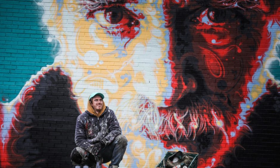 Street artist Michael Corr kneeling next to a mural of the singer Michael Marra in Dundee.