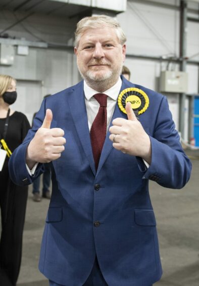 SNP MSP Angus Robertson wearing a SNP rosette and giving two thumbs up to the camera at an election count.