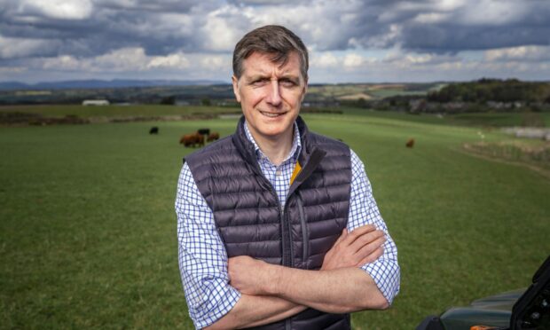 Simon Howie, arms folded in a field with cattle and hills behind him.