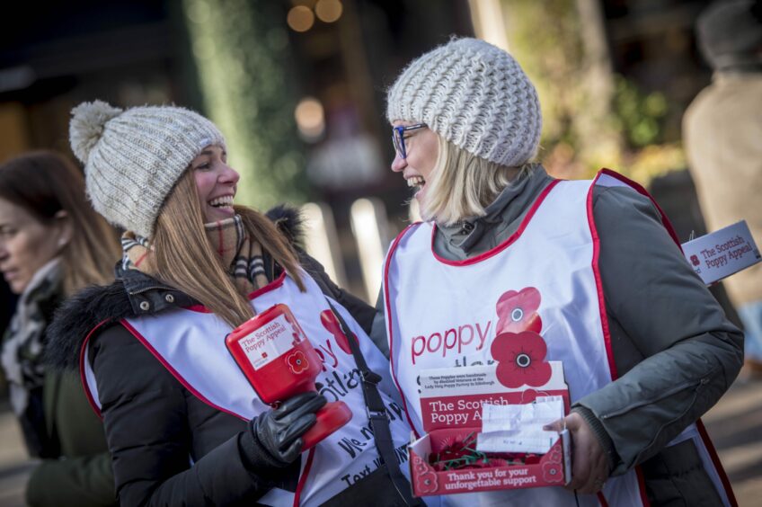 Two blonde women selling poppies for Poppyscotland are smiling and laughing while holding a cash box and a box of poppies. 