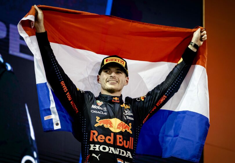 photo shows the F1 driver Max Verstappen on the winner's podium, holding a flag.
