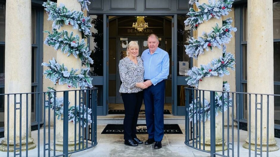 Margaret and Colin Smart at the entrance of the Dean Park Hotel in Kirkcaldy, holding hands in front of two columns wrapped with festive garlands. 