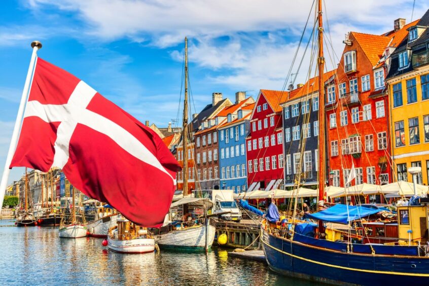 Photo shows a red and white Danish flag in front of the colourful old buildings and boats of Copenhagen's Nyhavn port.