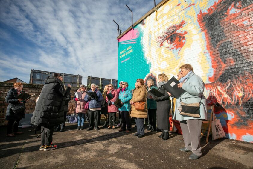 Photo shows a group of women in warm coats singing in front of a mural of the Dundee musician Michael Marra.