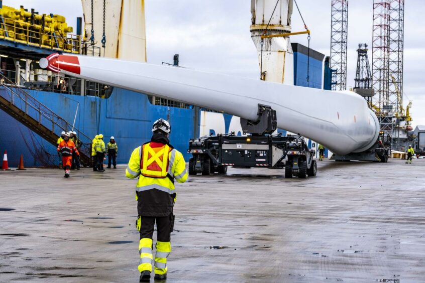 Photo shows large wind turbine blades on a trailer next to a ship at the Port of Dundee, with workers standing around.